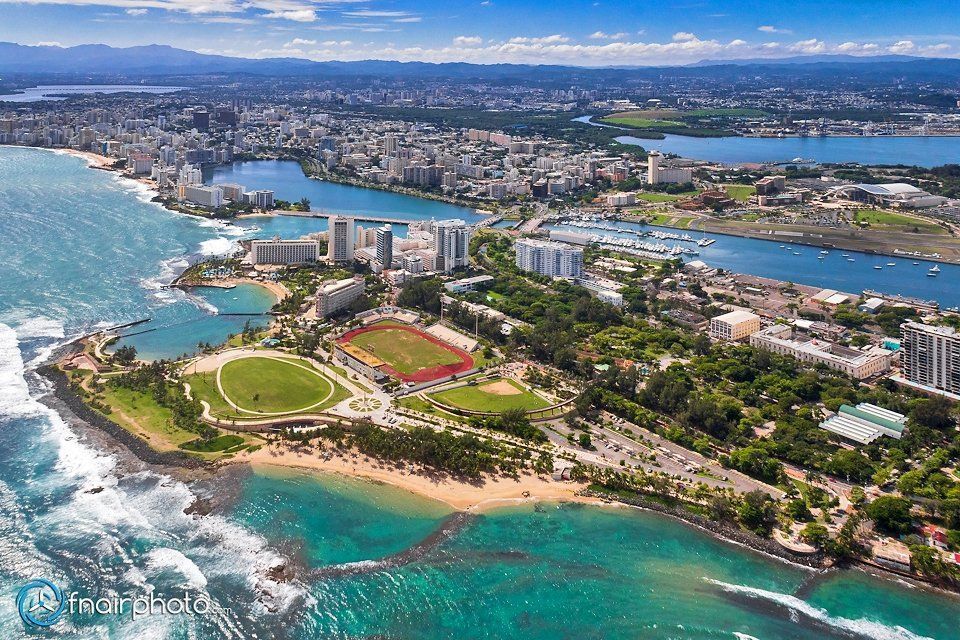 Aerial view of San Juan, Puerto Rico, with turquoise water, beaches, and a city skyline. A stadium is visible near the coastline.