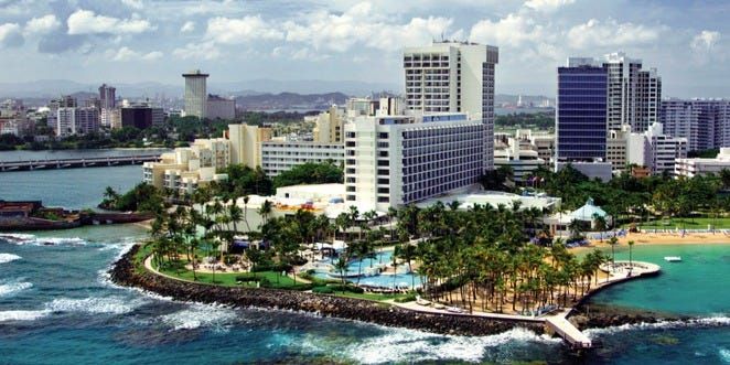 Aerial view of San Juan, Puerto Rico, showing hotels, a beach, and oceanfront buildings under a blue sky.