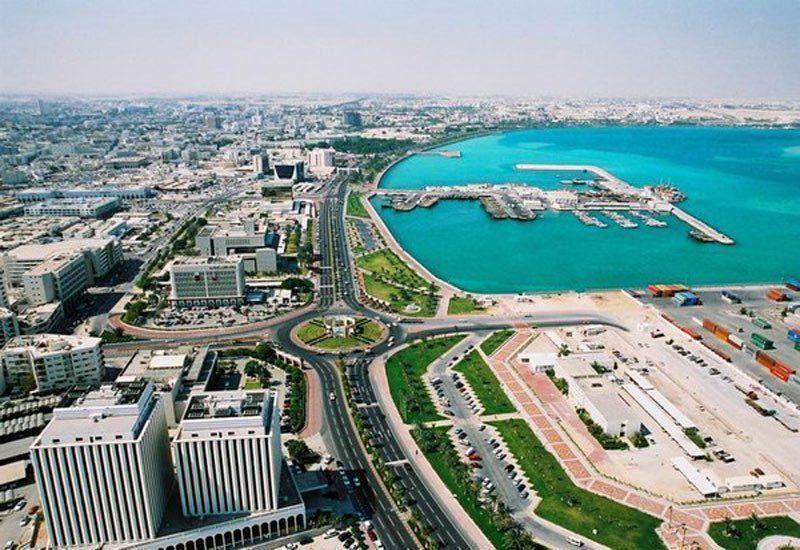 Aerial view of a coastal city with a harbor, turquoise water, roads, and buildings. A roundabout sits in the foreground.