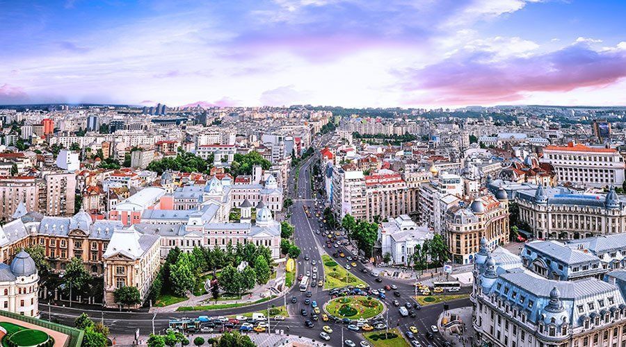 Aerial view of Bucharest, Romania, showcasing a wide boulevard lined with buildings, busy traffic, and colorful buildings under a pink and blue sky.