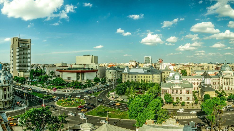 Cityscape with a tall modern building and classical architecture surrounding a green park and traffic circle under a blue sky.