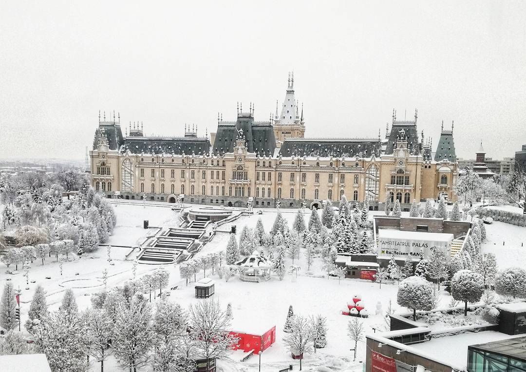 The Palace of Culture in Iasi, Romania, covered in snow. Large, ornate building with multiple towers, surrounded by snow-covered trees and a plaza.