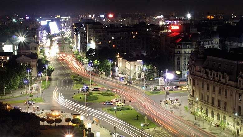 Nighttime view of a city street with moving car lights, buildings, and street lights illuminating the scene.