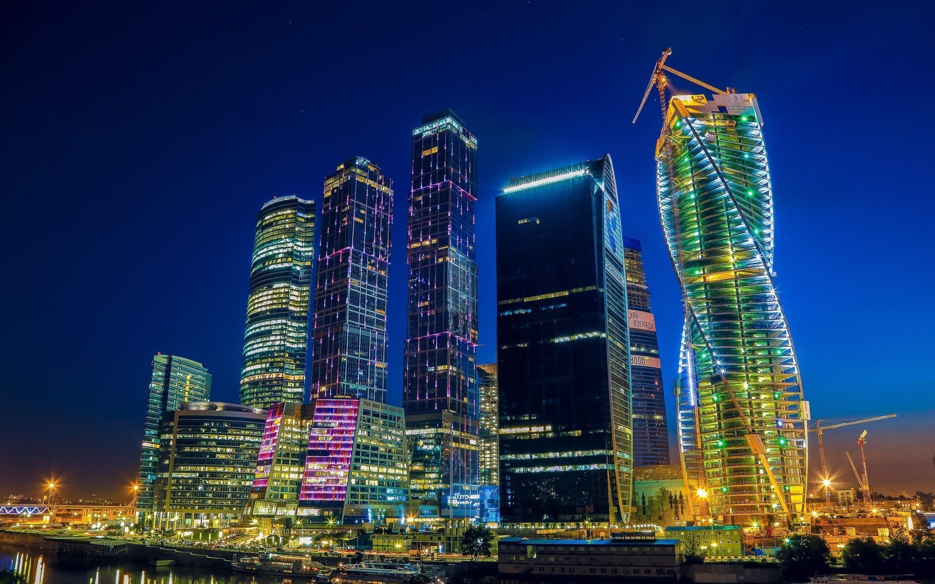 Skyscrapers illuminated at night against a deep blue sky; city lights reflect on water in the foreground.