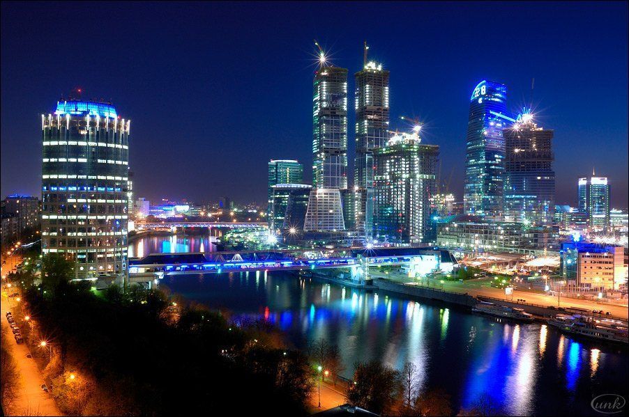 Nighttime cityscape reflecting lights on a river, with tall skyscrapers and bright illumination.