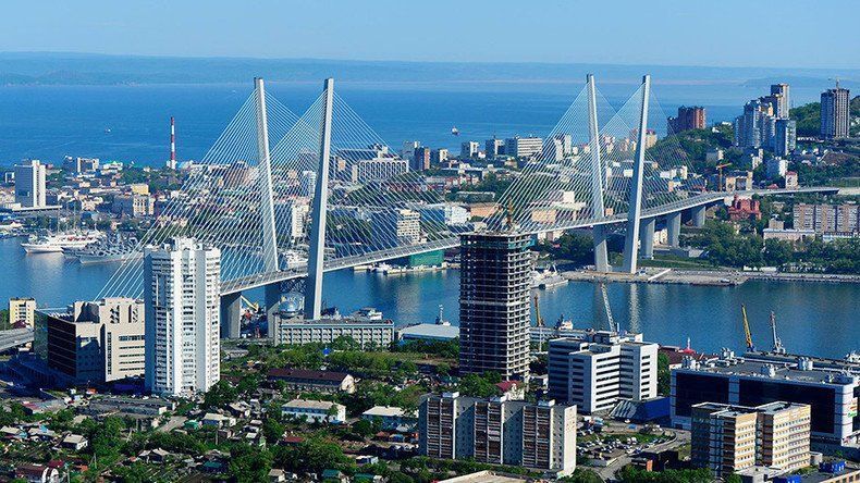 A panoramic view of Vladivostok, Russia, with a cable-stayed bridge spanning a harbor; buildings and sea in view.