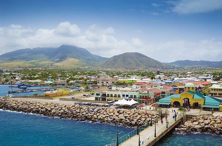 Coastal town with colorful buildings, pier, and mountains in the background under a partly cloudy blue sky.