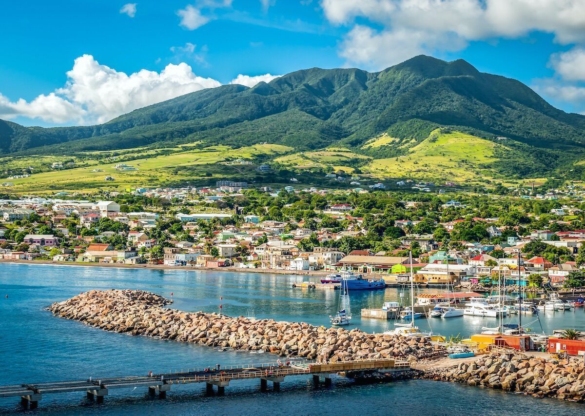 Coastal town with a mountain backdrop under a blue sky; boats in the harbor, buildings dotting the land.
