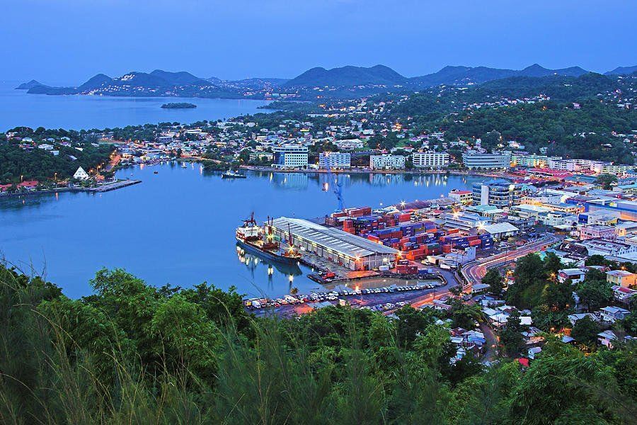 A harbor town at dusk, with buildings and container port. Hilly landscape with green trees. Blue sky and water.