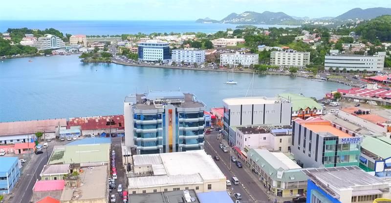 Aerial view of a coastal city with buildings along a bay, a blue-glass building in the center, and green hills in the background.
