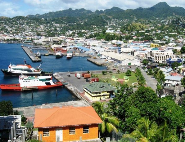 Coastal cityscape featuring a harbor with ships, docks, and buildings against a backdrop of green hills.