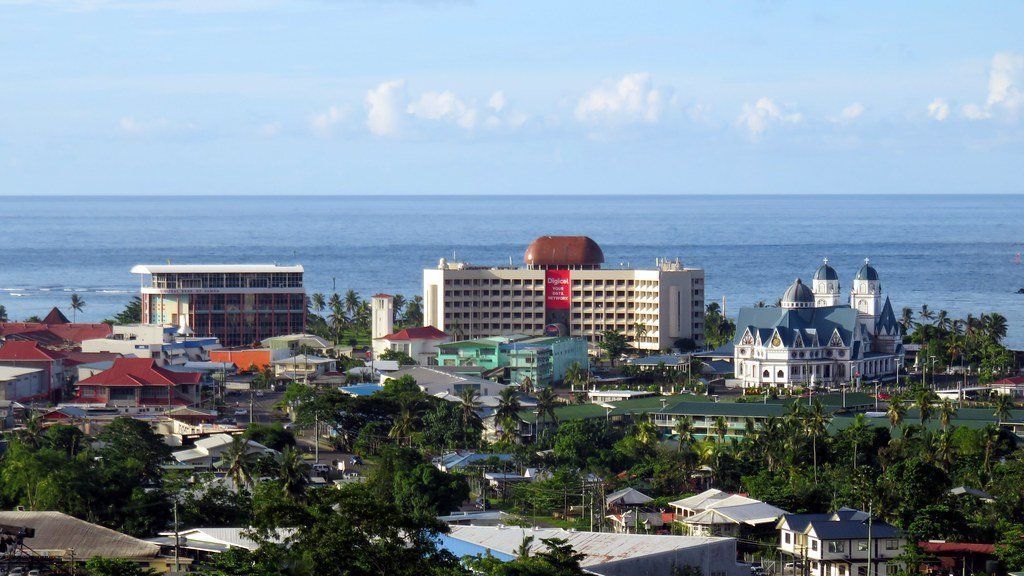 View of Apia, Samoa, with buildings of varying heights and styles near the coast. The sea is visible in the background.