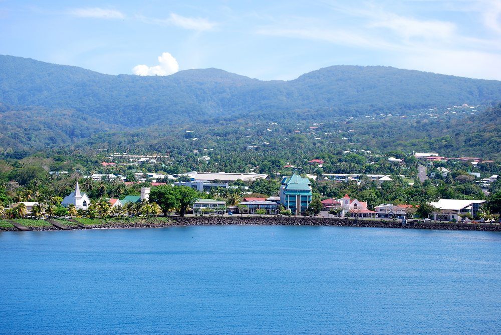 Coastal town nestled against green mountains, featuring a waterfront promenade, buildings, and blue sea under a clear sky.