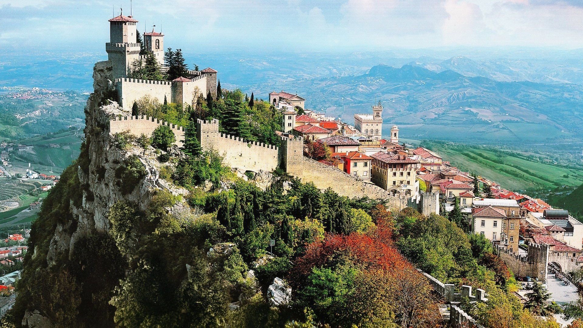 Medieval castle perched on a rocky mountaintop in San Marino, with surrounding buildings and a vast landscape in the background.