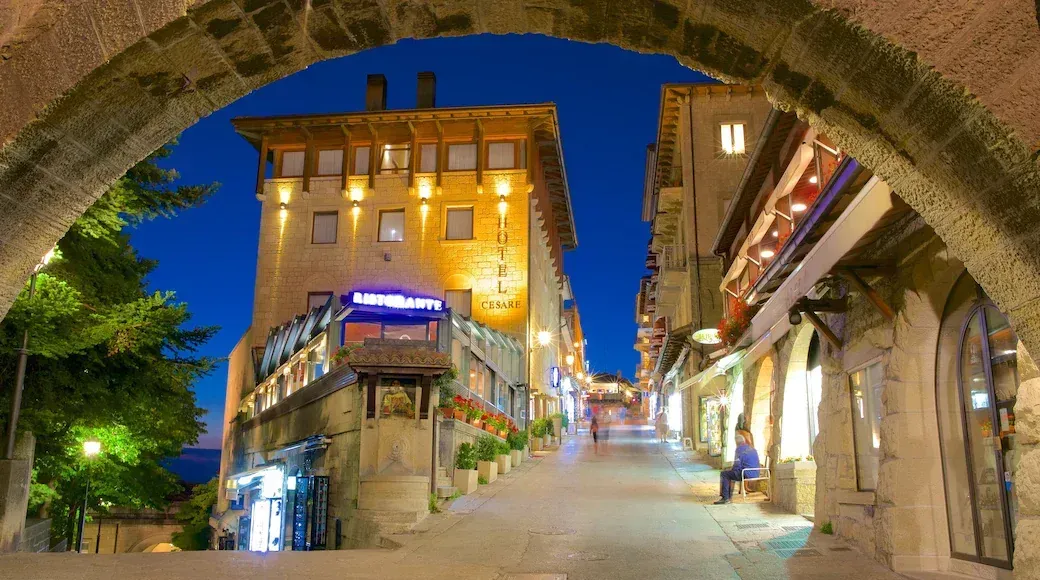 Stone archway framing a narrow, lit street at night, lined with shops and buildings. The main building is tall, with warm lighting.