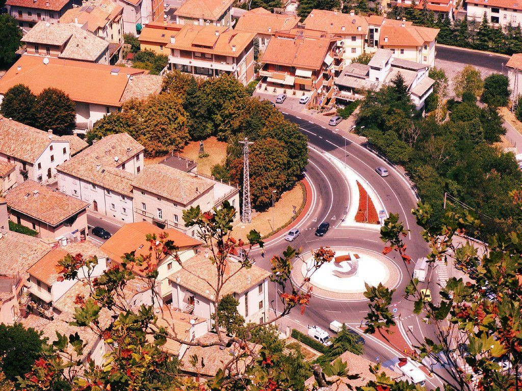 Aerial view of a European town with red-tiled roofs, a curved road, and a roundabout with a small sculpture.