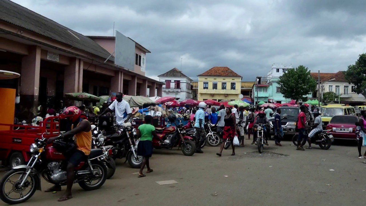 Busy market square in an African city. People gather around stalls, motorcycles, and cars under a cloudy sky.