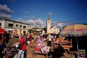 A busy market square with vendors and shoppers, a church with a tall steeple in the background under a blue sky.