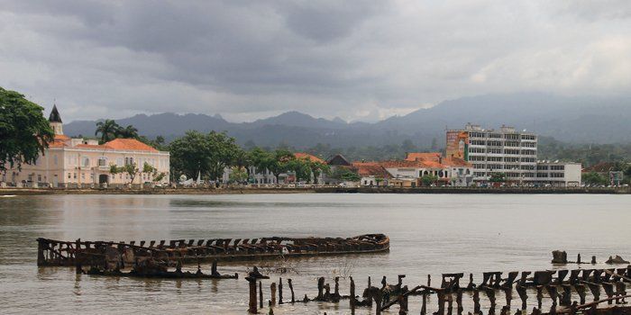 View of a city by the water under a cloudy sky, with buildings, mountains in the background, and the remains of a structure in the water.