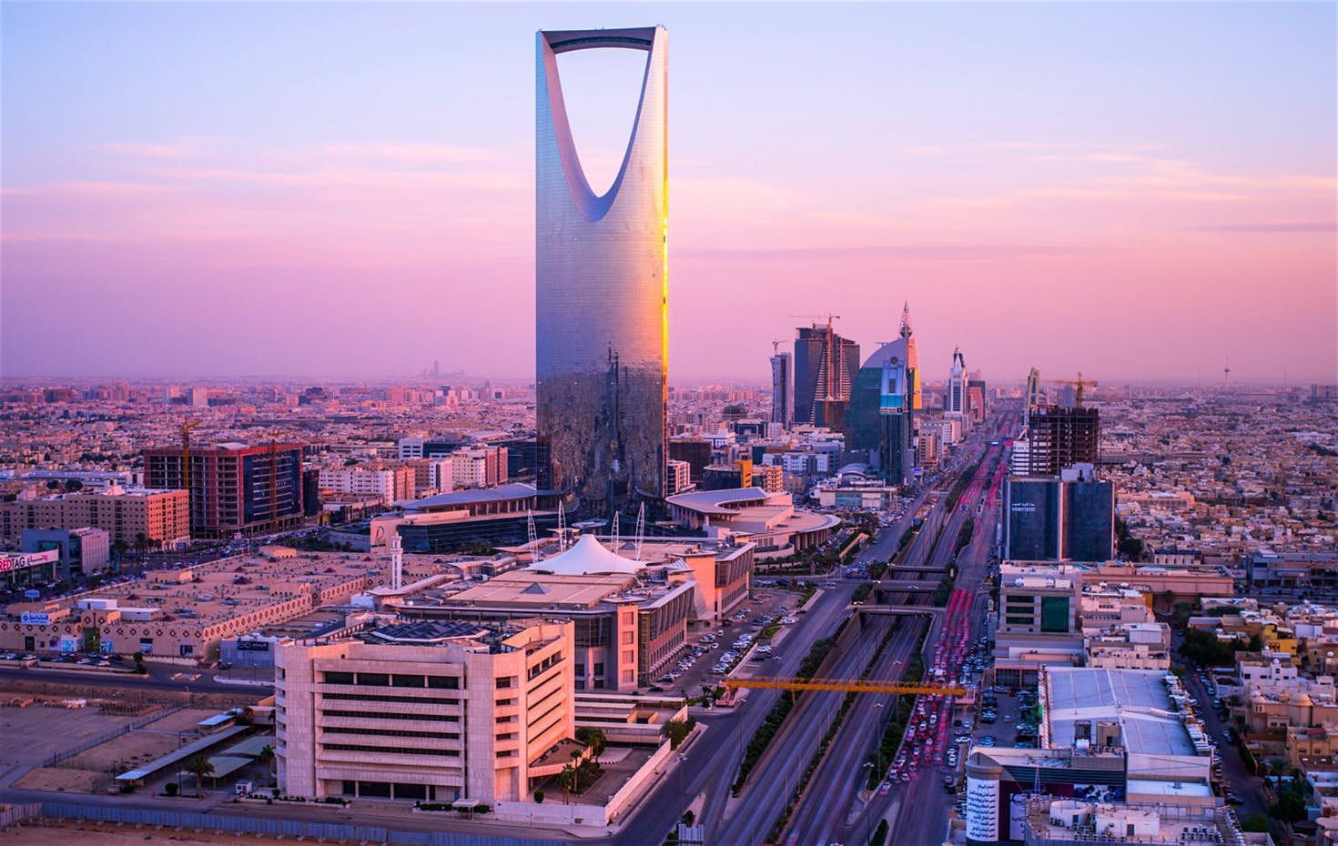 Aerial view of Riyadh, Saudi Arabia, dominated by the Al Faisaliah Center skyscraper at sunset with a pink and purple sky.