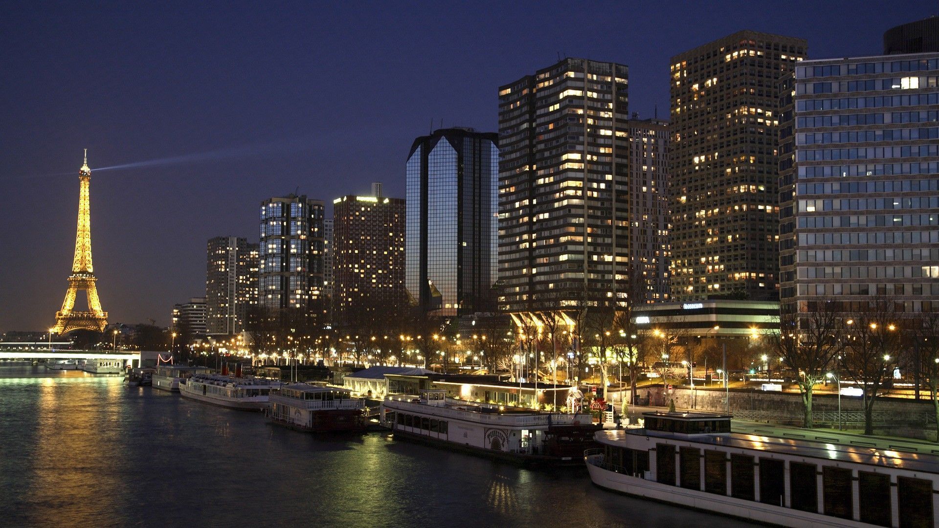 Eiffel Tower and illuminated cityscape at night, reflecting in the river Seine; boats line the water's edge.