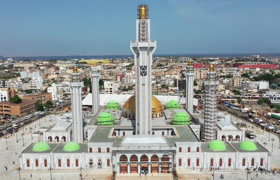 Aerial view of a large mosque in Dakar, Senegal, with a tall minaret and multiple domes. White building with green domes and a large paved courtyard.