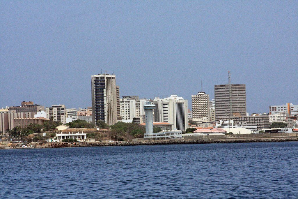 Skyline of Dakar, Senegal, viewed from the water. Several tall buildings, some with antennas, line the coast under a clear blue sky.