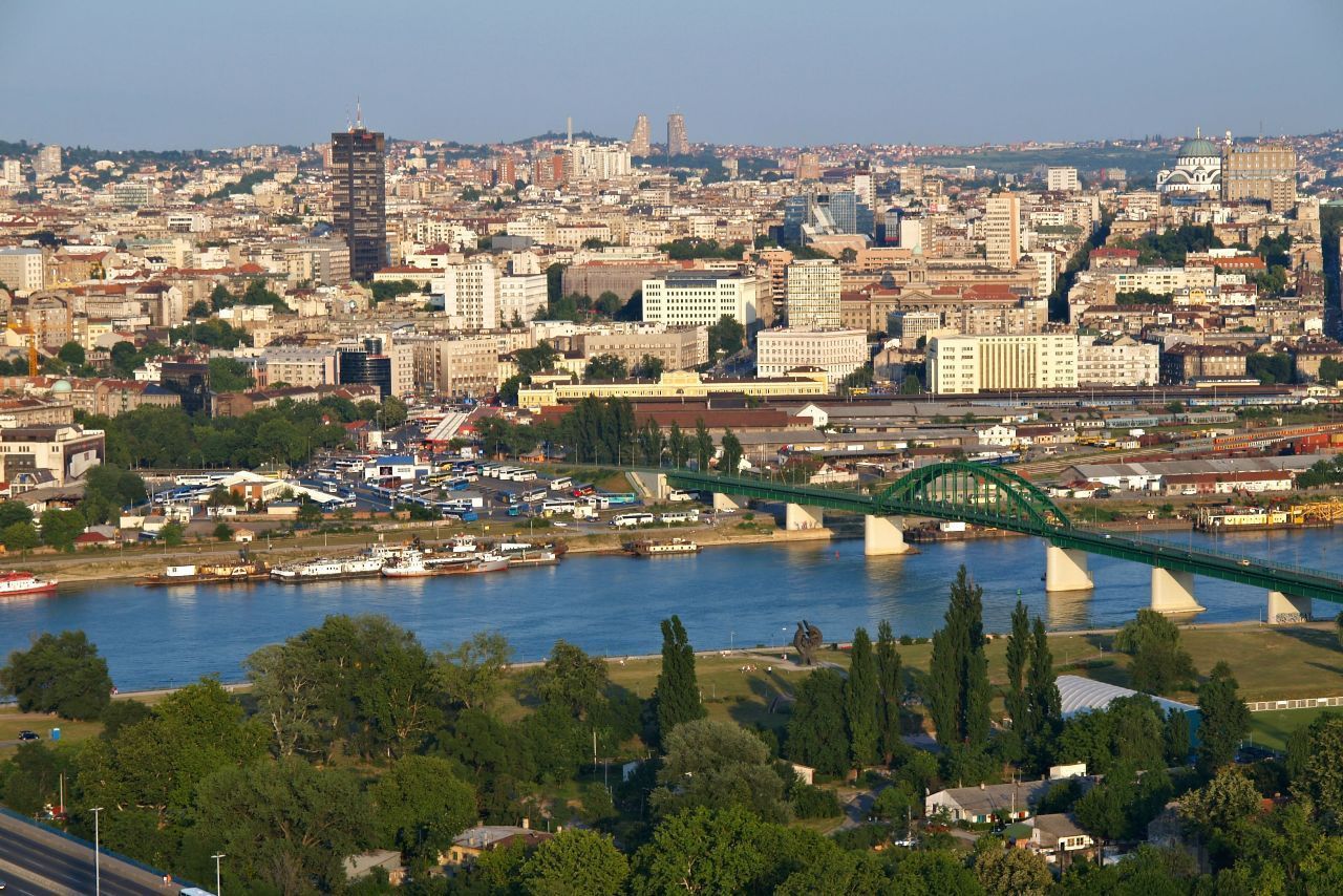 A cityscape view of Belgrade, Serbia, with a green bridge spanning a river, buildings, and a clear sky.