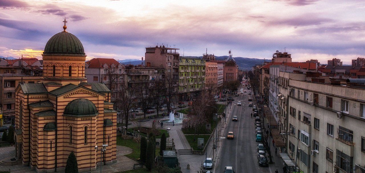 An aerial view of a city street at sunset with a large church on the left. Cars drive down the road as the sky is a blend of purples and oranges.