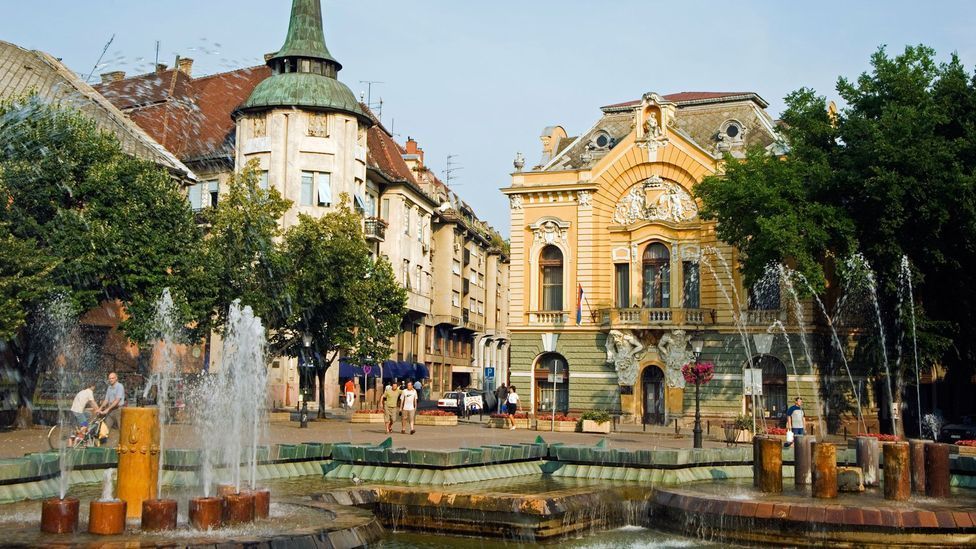 Fountain in a European town square with ornate buildings, trees, and pedestrians.