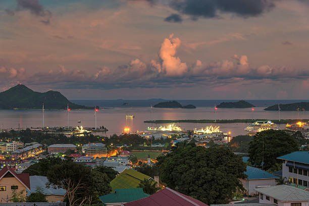 Panoramic view of a coastal city at dusk, with ships in the harbor and mountains in the background, under a colorful sky.