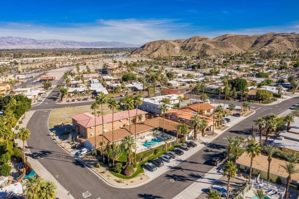 Aerial view of a neighborhood with palm trees and tan buildings. A pool and mountains are visible in the background under a blue sky.