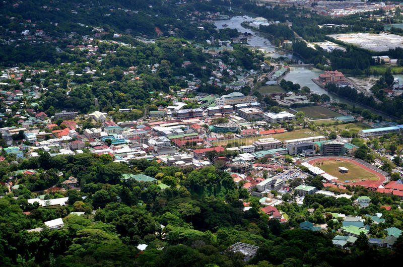 Aerial view of a coastal town, with buildings and vegetation. A river flows through the center, with a sports field visible to the right.