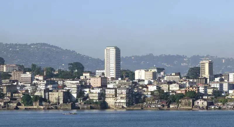 Skyline of a city on the water, with a tall white building as a focal point. Green hills and a blue sky complete the backdrop.