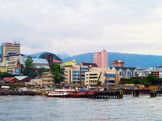 A coastal cityscape with buildings of various heights and styles along the water's edge. Boats are docked near a wooden pier under an overcast sky.