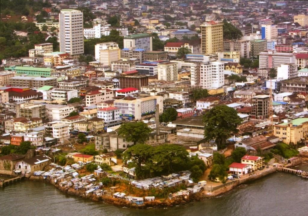 Aerial view of a city on the coast, likely in Africa. Buildings of various heights and colors are densely packed together.