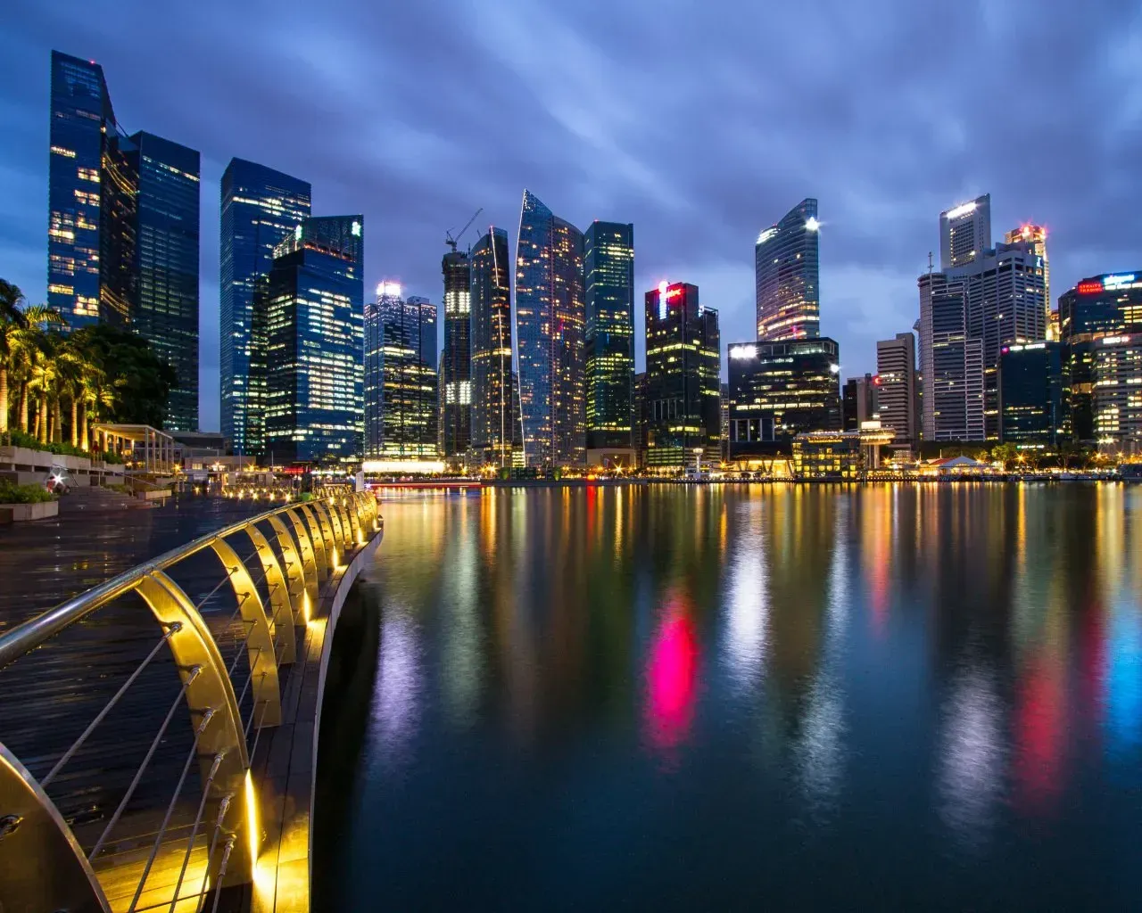 Nighttime cityscape with tall buildings reflecting in calm water, illuminated walkway in foreground.