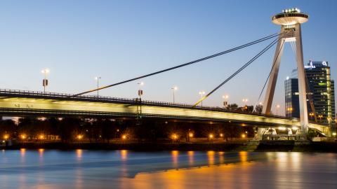 A lit cable-stayed bridge with a UFO-shaped observation deck crosses a dark river in Bratislava, Slovakia at dusk.