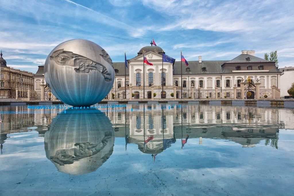 A large, reflective metal sphere sits in front of the presidential palace in Bratislava, Slovakia. The building is reflected in a shallow pool.