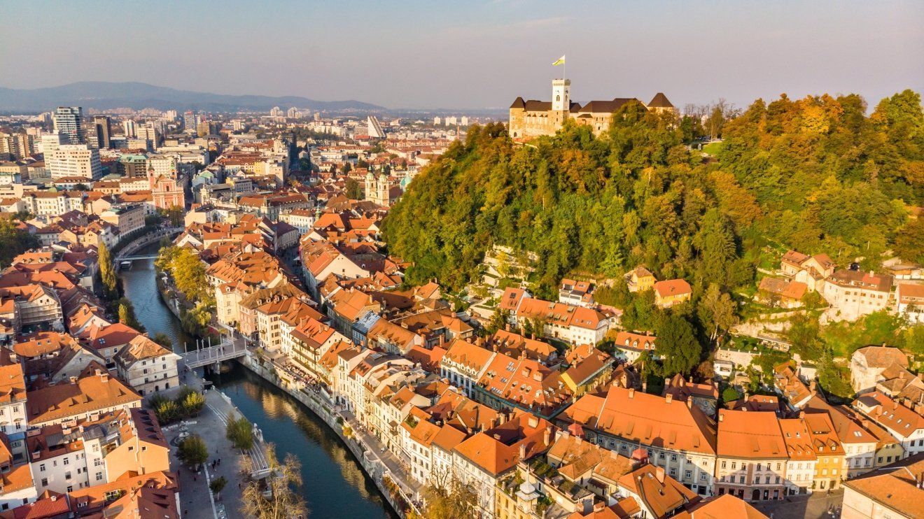 Aerial view of Ljubljana, Slovenia, with the castle atop a hill, the Ljubljanica River winding through the city, and orange rooftops.