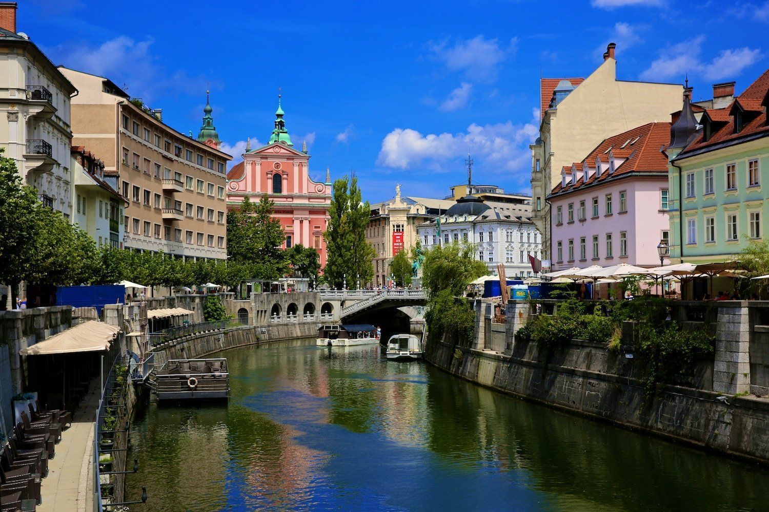 A canal flows through a European city with colorful buildings. Pink church with two spires in the background.