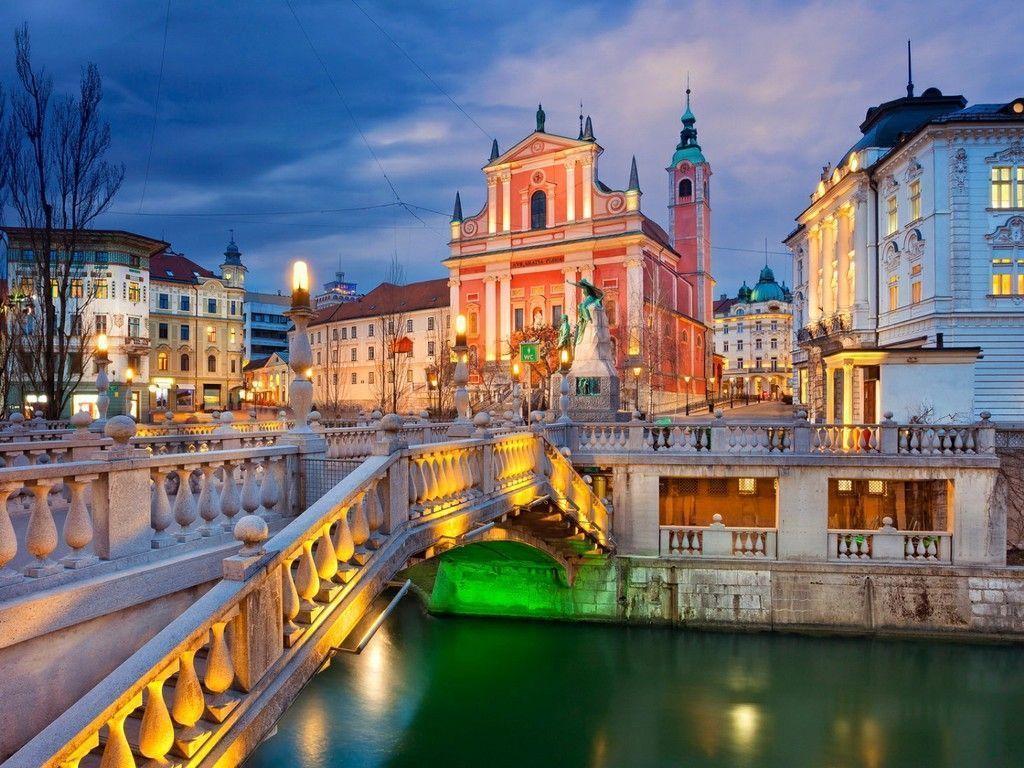 Ljubljana, Slovenia at dusk: illuminated Triple Bridge over the Ljubljanica River with a baroque church in the background.
