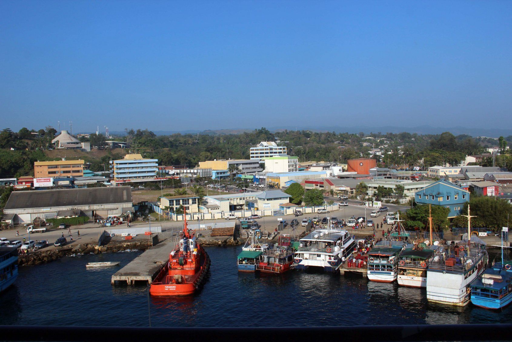 Harbor view with several boats docked near a town with buildings and trees under a clear blue sky.