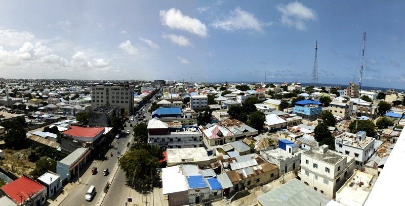 Panoramic view of Mogadishu, Somalia, under a blue sky with scattered clouds. The cityscape features buildings of varying heights and colors.