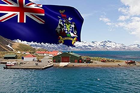 A coastal scene featuring a flag of South Georgia and the South Sandwich Islands, buildings on a pier, mountains, and a body of water under a blue sky.