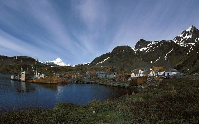 A harbor town nestled beneath snowy mountains; buildings line the shore, with a blue sky streaked with clouds.