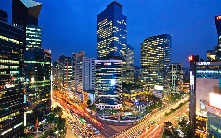 Night view of a modern city with brightly lit skyscrapers and car light trails along a road.