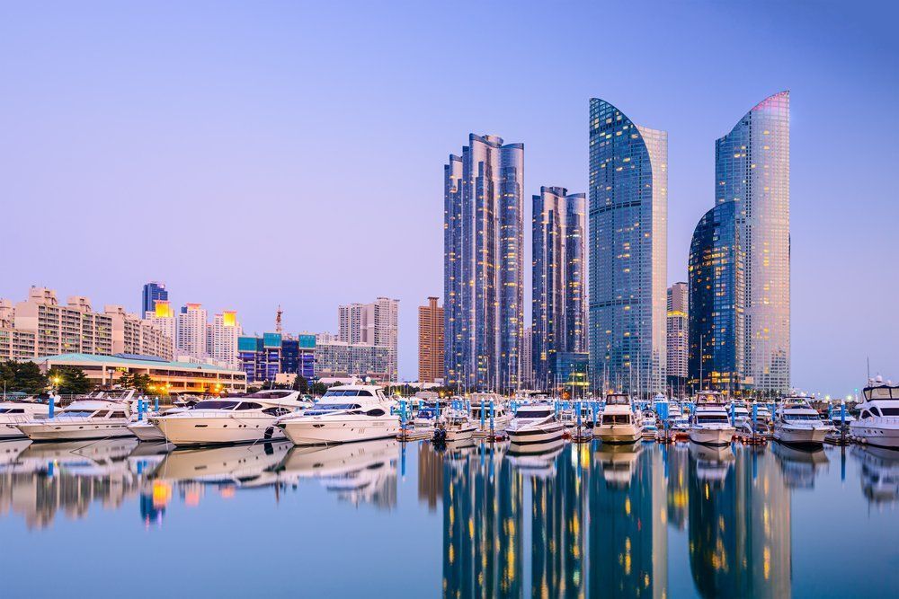 Marina with boats in front of modern skyscrapers at dusk, reflecting in the water.