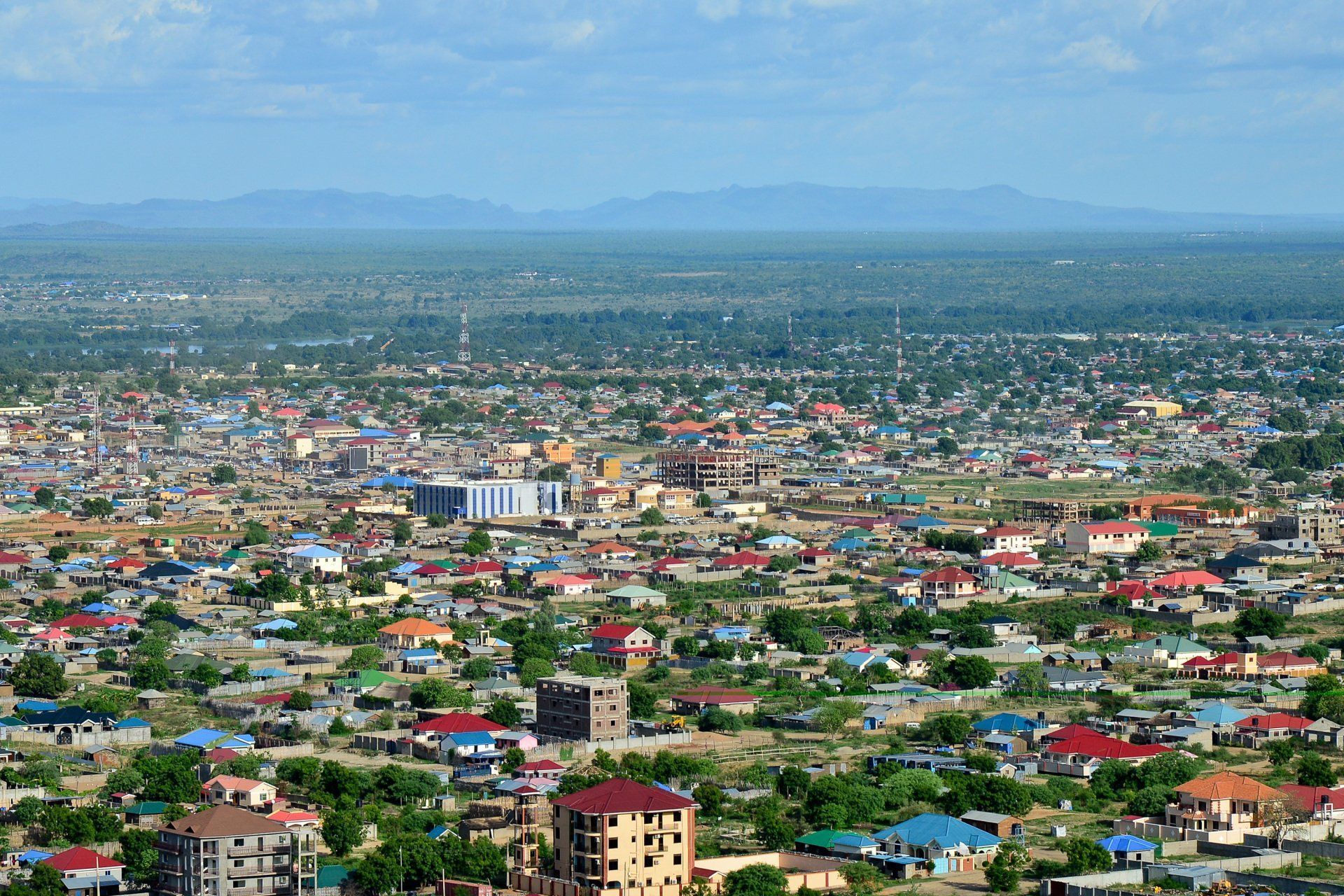 Aerial view of a densely populated town with buildings and trees. Distant mountains are visible under a blue sky.