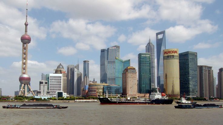 Shanghai skyline with the Oriental Pearl Tower in the foreground, several skyscrapers behind it, and cargo ships on the river below a blue sky.